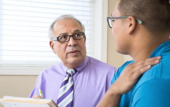 A male doctor wearing glasses, a purple shirt, and a striped tie looks intently at a patient in a blue shirt during an office consultation.